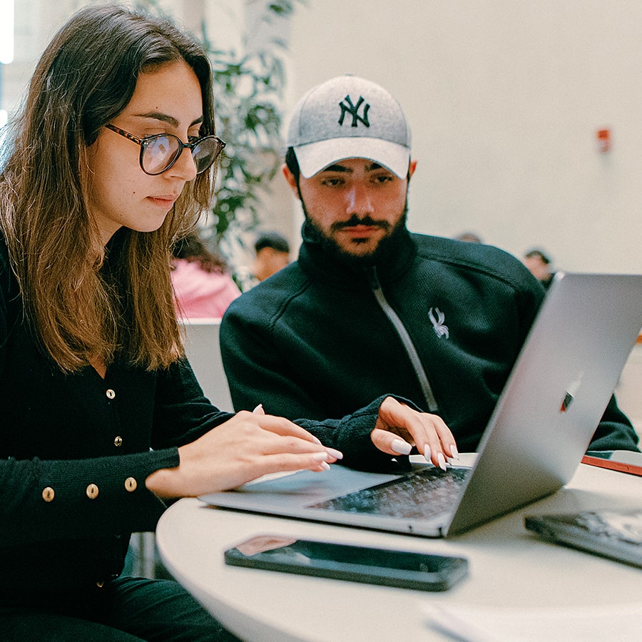 Two students one in baseball hat look over laptop in campus center.