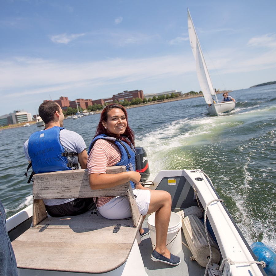 Two students on a motorboat with students on sailboat and campus in background.