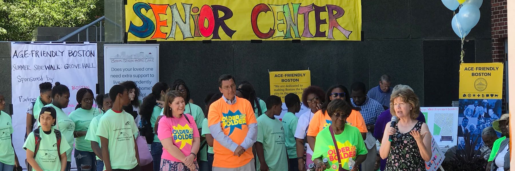 Crowd at event in front of the Grove Hall Senior Center under a banner.